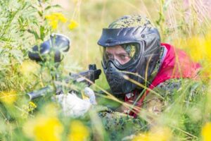 Paintball sport player in protective uniform and mask playing with gun outdoors and sneaking in grass