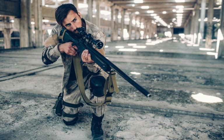 One dark-haired man is standing in long hangar by himself. he has put one knee down on the ground and holding rifle in hands